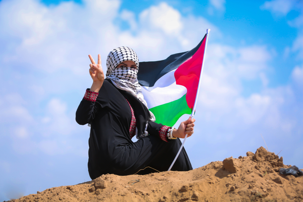 Close up of a woman holding a Palestinian flag on top on a mound in holding her fingers in a sign for victory